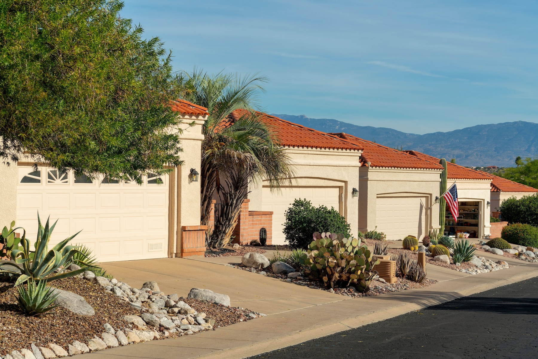 Row of modern adobe style one story houses in desert community of arizona with visible garage doors and front yards. In afternoon sun with red roof tiles and gable design with driveways